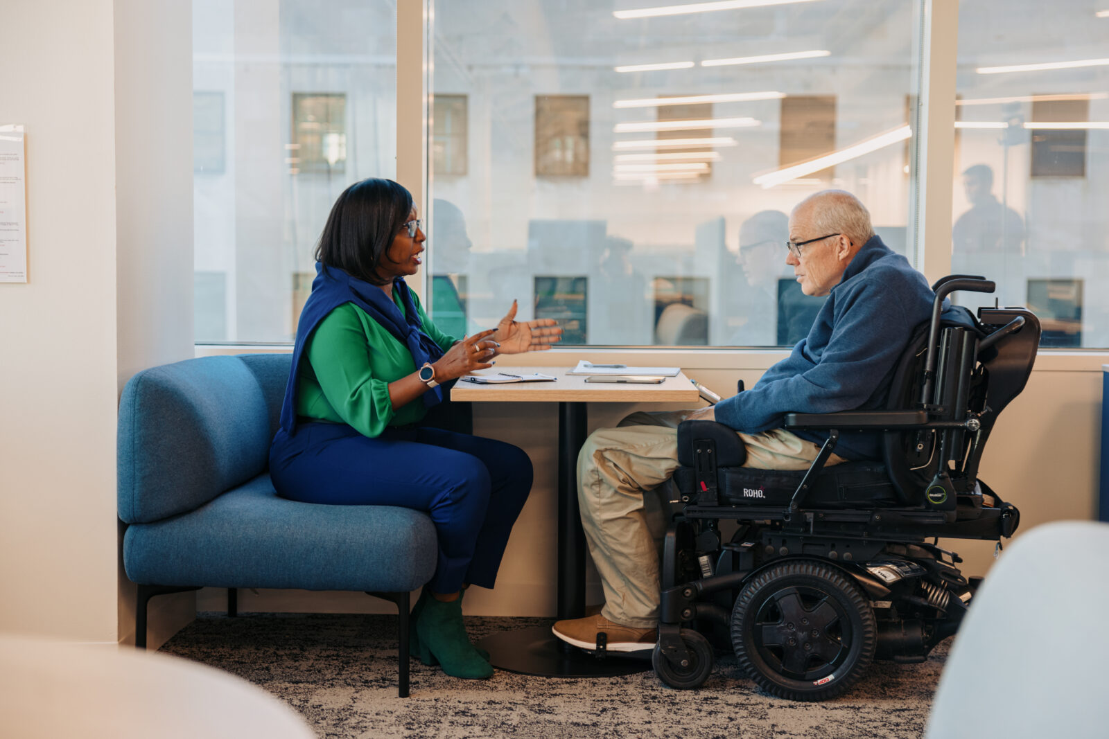 Two disabled people converse and sit across from each other at a table with pens and notebooks on top of it. One uses a power wheelchair; a tablet is on the table in front of them. Photo by Jordan Nicholson for Disability:IN.
